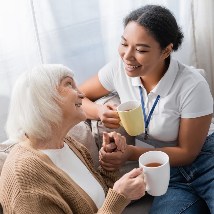 cheerful multiracial social worker chatting with senior woman while having tea in living room